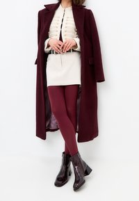 Burgundy coat over a beige top with gold buttons, white mini skirt, maroon tights, and black shiny ankle boots with a block heel.