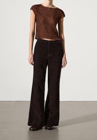 Ruskea neulottu lyhythihainen crop-top, yhdistetty tummanruskeisiin corduroypöksyihin, seisoo vaaleanharmaalla pinnalla.