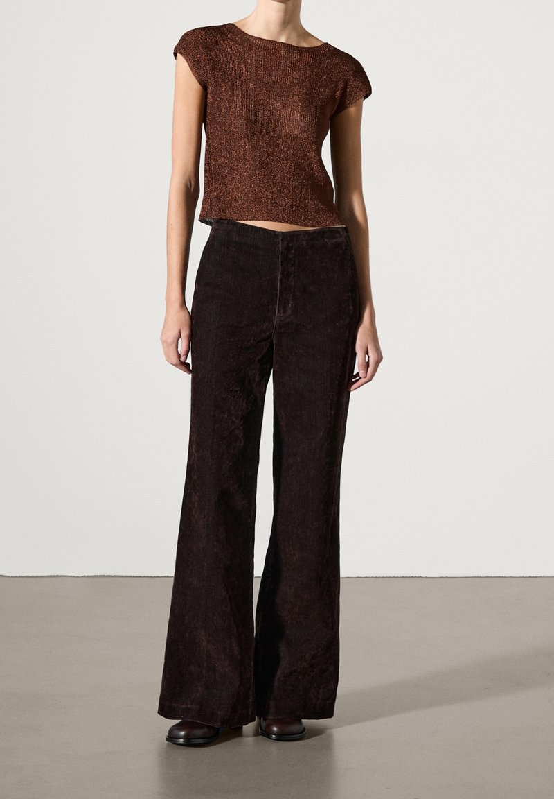 Ruskea neulottu lyhythihainen crop-top, yhdistetty tummanruskeisiin corduroypöksyihin, seisoo vaaleanharmaalla pinnalla.