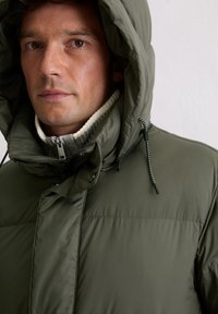 Olivengroen donsjack met een grote capuchon, voorzien van een ritssluiting aan de voorkant en trekkoorden als accent. De binnenkant heeft een geribbelde, lichtkleurige kraag.