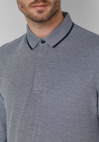 Mann in einem grauen Poloshirt mit einem strukturierten Kragen und einem einzigen Knopf, der sichtbar einen Teil seines Bartes und Halses zeigt.