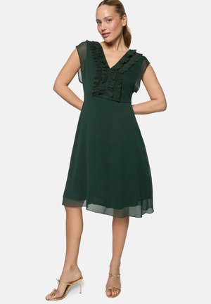 Frau trägt ein dunkelgrünes knielanges Kleid mit gerafftem V-Ausschnitt und transparenten kurzen Ärmeln, kombiniert mit goldenen Sandalen mit hohem Absatz.