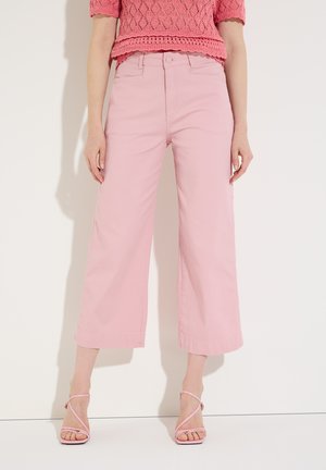 Femme portant un pantalon rose taille haute à jambes larges, des talons à brides roses et un haut rose texturé à manches courtes, debout contre un mur blanc.