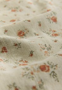 Jasnobeżowa tkanina z kwiatowym wzorem w stonowanych odcieniach czerwieni i zieleni. Teksturowana powierzchnia z widocznym skośnym splotem i rozrzuconymi małymi kwiatkami.