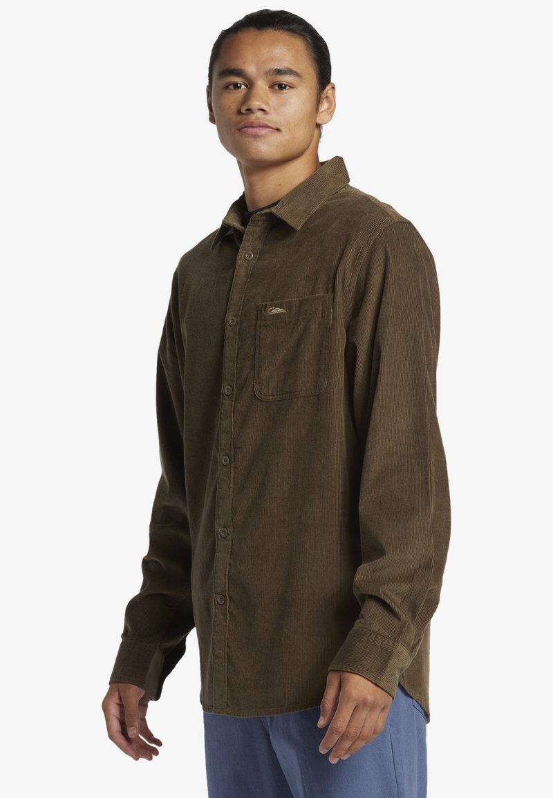 Camicia button-up in corduroy marrone con tasca frontale e logo ricamato. Maniche lunghe con polsini a bottone, orlo dritto, texture morbida.