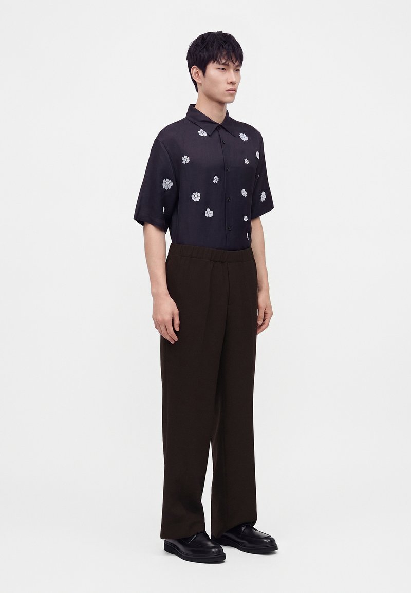 Hombre con camisa negra de manga corta y botones con estampados florales blancos, pantalones anchos oscuros y zapatos negros, de pie frente a un fondo liso.