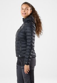 Donna con capelli lunghi e ricci che indossa una giacca imbottita nera e pantaloni scuri da esterno, in piedi di profilo su uno sfondo chiaro e uniforme.