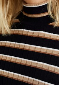 Acercamiento a un suéter de cuello alto negro con canalé, con franjas horizontales beige, marrón y blancas, llevado por una persona con cabello rubio.