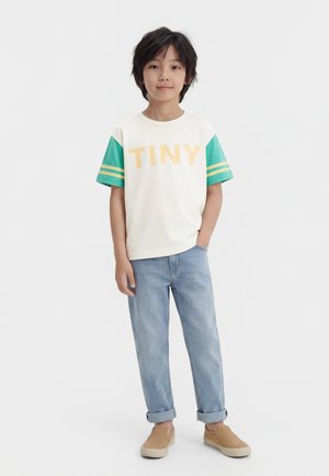 Otrok stoji in nosi belo majico z zelenimi rokavi in napisom "TINY", svetlo modre zvite kavbojke ter rjave obutev brez vezalk.