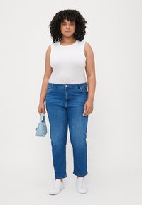 Top branco sem mangas, calças de ganga azuis, sapatilhas brancas e uma pequena mala azul. As calças têm um corte reto e estão ligeiramente desgastadas na bainha.