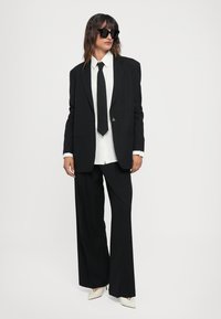 Black tailored suit featuring a structured blazer, wide-leg trousers, white shirt underneath, and a black tie. Accessories include large black sunglasses.