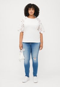 A woman with curly hair wears a white top with lace sleeves and blue ripped jeans, holding a small white bag, standing against a plain background.