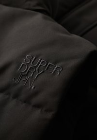 Zwarte stof met gestikte tekst "SUPER DRY JPN." op gewatteerd materiaal, waarschijnlijk onderdeel van een jas of mantel.