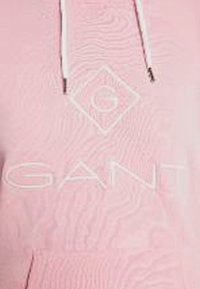 Sudadera de color rosa claro con una superficie texturizada, que presenta un logo blanco y detalles en blanco. Incluye un bolsillo frontal y cordones ajustables en blanco.