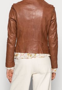 Hnědá kožená bunda s přiléhavým střihem, která má hladký povrch a švy, navrstvená přes květinovou blůzu s volány.