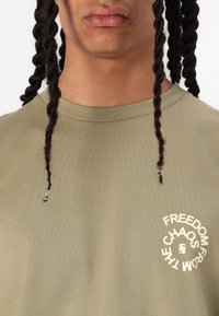 Olive green short-sleeve t-shirt featuring a circular beige graphic with the text "FREEDOM FROM CHAOS." Smooth fabric with a crew neckline.