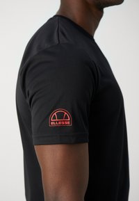 Camiseta negra de manga corta hecha de tejido suave, con un parche de logo rojo en el hombro izquierdo. Diseño clásico de cuello redondo.