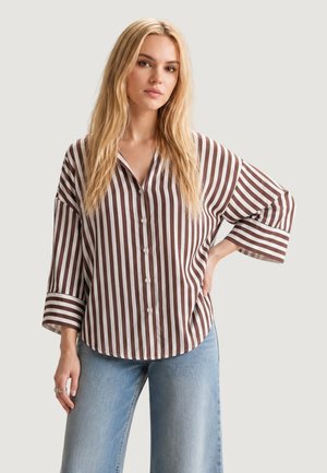 Vrouw draagt een losse bruin-wit verticaal gestreepte blouse met wijde mouwen en lichtblauwe jeans, staand met hand in de zij.