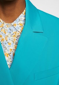 Blazer turchese con rever affilati, abbinato a una camicia azzurro chiaro a motivo floreale con accenti arancioni e gialli.