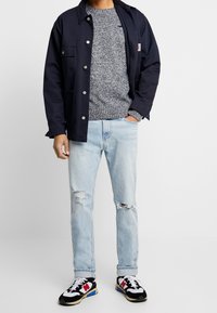 Man med marinblå jacka, grå stickad tröja, ljusa slitna jeans och svarta, vita, röda och blå sneakers står med ena handen i fickan.