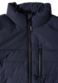 Marinblå isolerad jacka med hög krage, fullt dragkedja fram och en ficka på bröstet med dragkedja. Har en quiltad textur och en slank design.