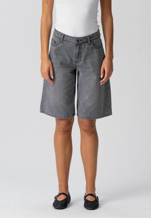 Osoba ubrana w szare dżinsowe szorty do kolan, biały bezrękawnik i czarne płaskie buty, stojąca na jednolitym tle.