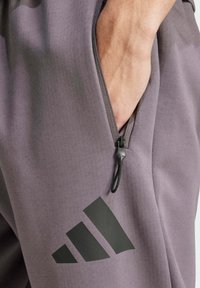 Gray athletic pants feature a zippered pocket with a black drawstring, and a prominent black Adidas logo. Smooth fabric with a soft texture.