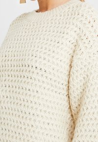 Cremefarbener gestrickter Pullover mit einem strukturierten Muster, rundem Ausschnitt und langen Ärmeln. Das Material wirkt weich und atmungsaktiv.