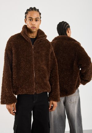 Due persone con capelli intrecciati che indossano giacche zip-up marroni e pelose, una di fronte e l'altra che mostra il retro della giacca.