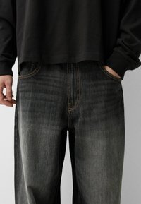 Tummat farkut suoran istuvuuden kanssa, haalistunut etuosa ja erottuva oranssi tikkaus taskuissa ja saumoissa. Yhdistetty löysään mustaan paitaan.