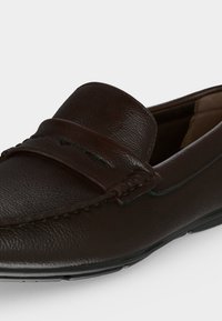 Gros plan sur un mocassin en cuir marron foncé avec des coutures apparentes et un design classique à fente pour pièce sur le dessus.