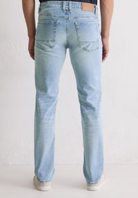 Personne portant un jean droit bleu clair et des baskets blanches, se tenant devant un mur blanc uni.