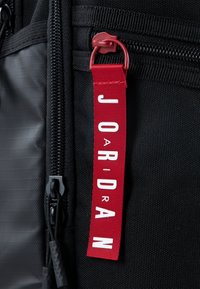 Fekete hátizsák, texturált anyagból, piros címkével, fehér betűkkel, melyen a "J O R D A N" felirat olvasható, és piros zipzárhúzóval.