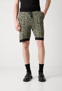 Pantalones cortos de patrón negro y verde con cintura ajustable y puños negros, hechos de un material suave. Combinados con calcetines oscuros y zapatos.