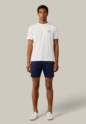 Junger Mann steht, trägt ein weißes kurzärmeliges T-Shirt mit Logo auf der Brust, marineblaue Shorts mit Logo und weiße Sneakers vor einem einfarbigen Hintergrund.
