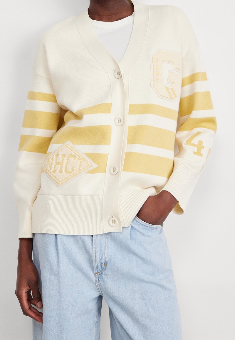 Cardigan crema con righe gialle, scollatura a V e chiusura con bottoni. Presenta toppe geometriche "G" e "WHCT" con polsini e orlo a coste.