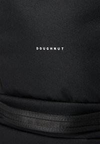 Crni platneni ruksak s teksturiranom površinom, s bijelim logotipom "DOUGHNUT" i crnom trakom koja prolazi preko tijela.
