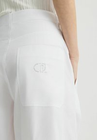 Weiße Baumwollhose mit lockerer Passform, ausgestattet mit einer Seitentasche und einem metallischen Logo-Detail auf der Gesäßtasche. Glatte Textur und klare Linien.