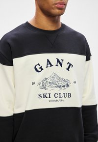 Muž v černobílém svršku s nápisem "GANT Ski Club Colorado, USA" a grafikou horské krajiny a lyžaře na hrudi.