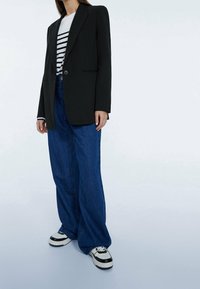 Černý oversized blazer, tričko s bílými a černými proužky, volné modré džíny a černo-bílé tenisky. Hladká tkanina a strukturovaný design.