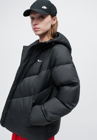 Crna zimska jakna s kapuljačom, koja ima prednji zip, elastične manžete i mali bijeli Nike logo na prsima. Glatka tekstura.