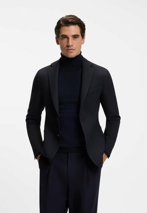 Man draagt een donkerblauw colbert over een zwarte coltrui met bijpassende donkere broek, staand met handen in de zakken tegen een witte achtergrond.