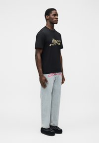 Μαύρο βαμβακερό t-shirt με γραφικό σχέδιο μπροστά, σε συνδυασμό με ανοιχτό μπλε τζιν με ίσια γραμμή. Το μοντέλο φοράει μαύρα παπούτσια slip-on.