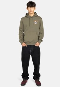 Sweatshirt olive avec une poche avant et un design graphique, associé à un jean noir ample et des chaussures bordeaux. Tenue simple et décontractée.