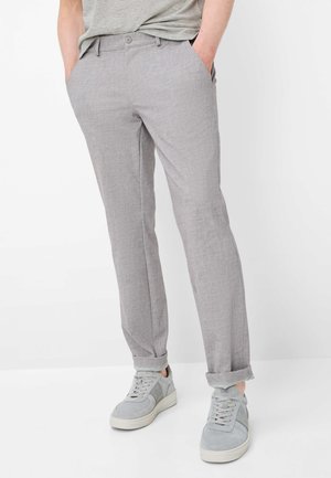 Person trägt hellgraue, schlank geschnittene Hose mit hochgekrempeltem Saum und passende graue Sneakers, steht mit den Händen in den Taschen.