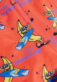Punainen kangas, jossa on leikkisä banaani piirretty aurinkolaseissa ja surffilaudan kanssa. Teksti kuuluu "TOP BANANA" tähtikoristeiden kera.