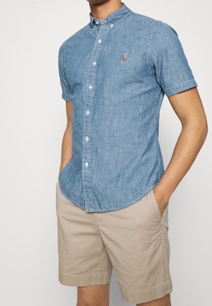 Homme portant une chemise à manches courtes bleu clair boutonnée et un short beige, les mains dans les poches, sur fond blanc.