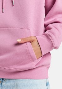 Růžová mikina vyrobená z jemné látky, s přední kapsou a žebrovanými manžetami. Design zahrnuje šňůrky se stříbrnými konci.