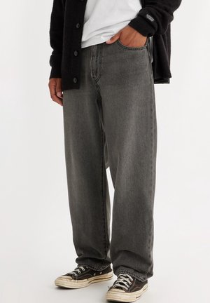 Person in lockeren grauen Jeans, schwarzer Strickjacke, weißem Hemd und schwarz-weißen Turnschuhen, Hand in der Tasche, steht vor weißem Hintergrund.