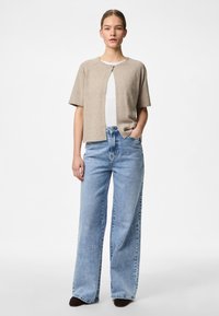 Cardigan bege de manga curta com decote redondo e fecho de botão, combinado com jeans de perna larga azul claro e sapatos castanhos escuros.